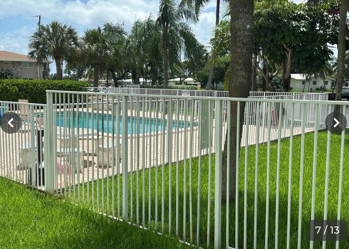 A white metal fence surrounds a swimming pool area with palm trees in the background — 151 SE 6th Avenue # 12, Pompano Beach FL 33060, POMPANO BEACH, FL