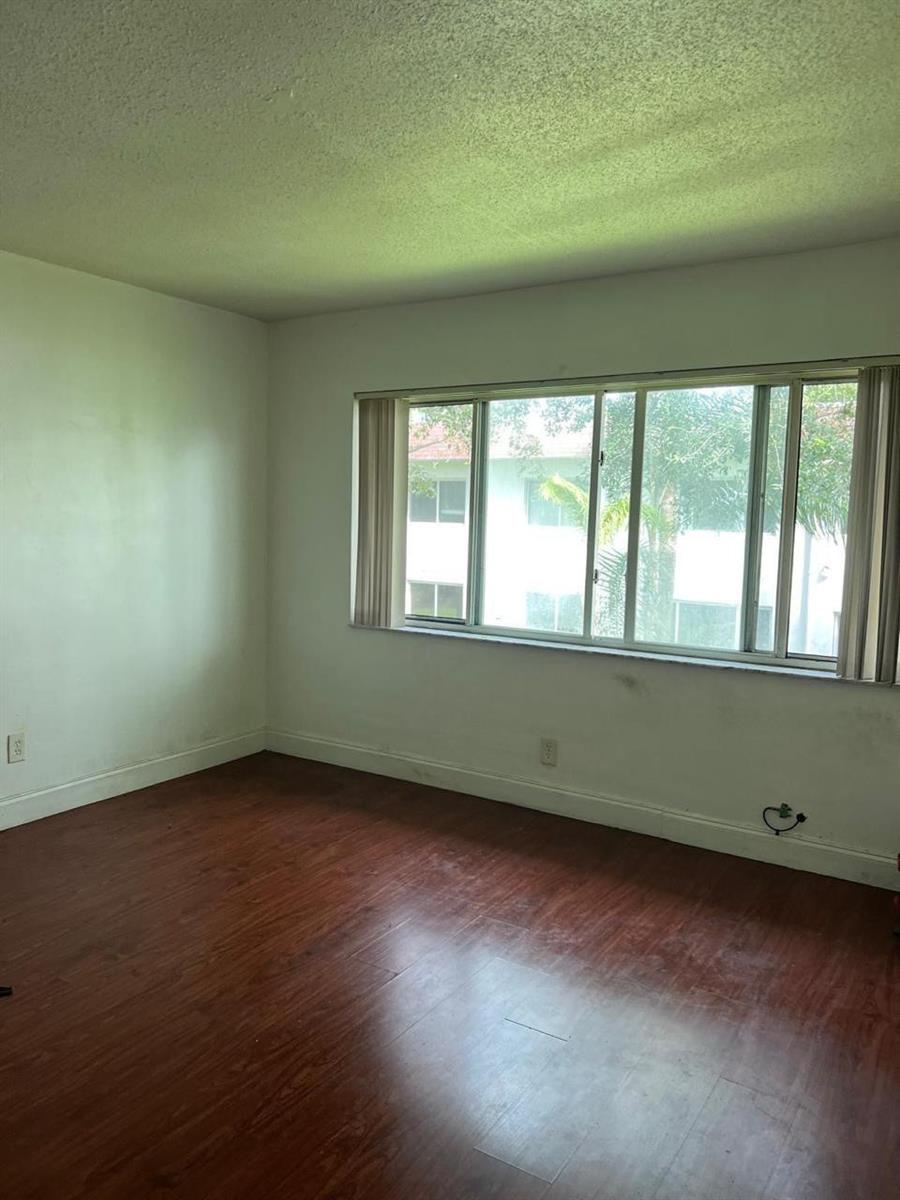 An empty room with wood-look flooring, white walls, and a large window overlooking trees — 151 SE 6th Avenue # 12, Pompano Beach FL 33060, POMPANO BEACH, FL