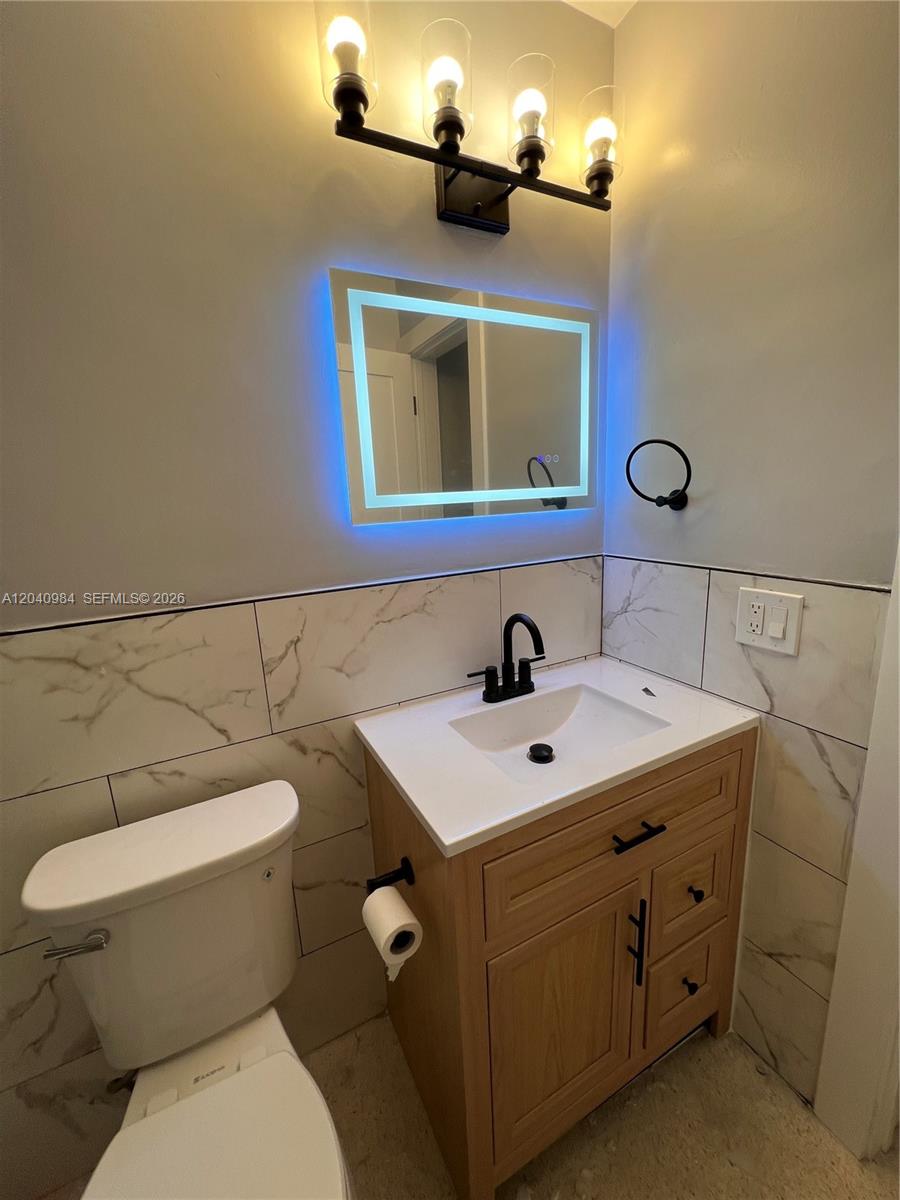 A bathroom features a wood vanity, a backlit mirror, and white marble-patterned wall tiles — 1140 NE 24th Ave # 1, Pompano Beach FL 33062, POMPANO BEACH, FL
