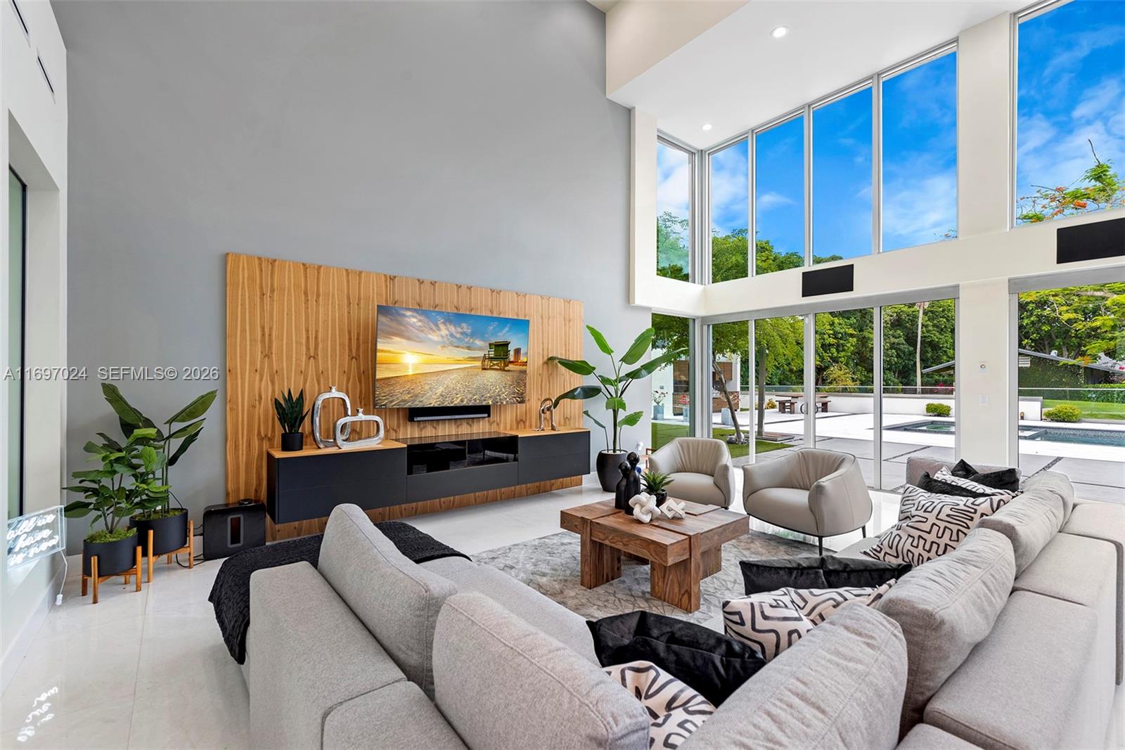 A modern living room features a wood-paneled media wall, high ceilings, and floor-to-ceiling windows — 14305 Stirling Rd, Southwest Ranches FL 33330, SOUTHWEST RANCHES, FL