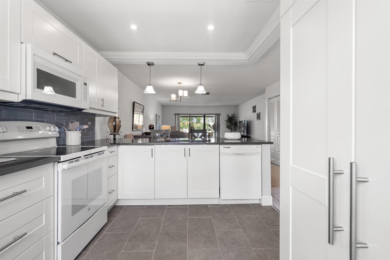 A white kitchen with dark countertops, grey tile flooring, and an open view to the living area — 21218 Lago Circle # F, Boca Raton FL 33433, BOCA RATON, FL