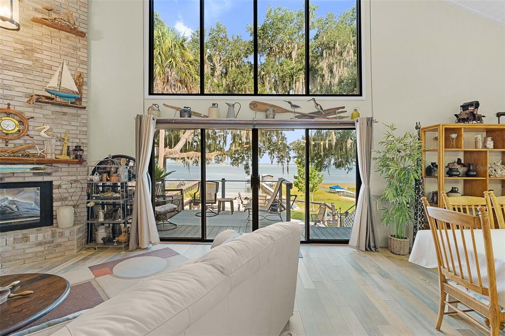A living room with a brick fireplace, vaulted ceilings, and large windows overlooking a deck — 15562 SE 138TH TERRACE, WEIRSDALE, FL