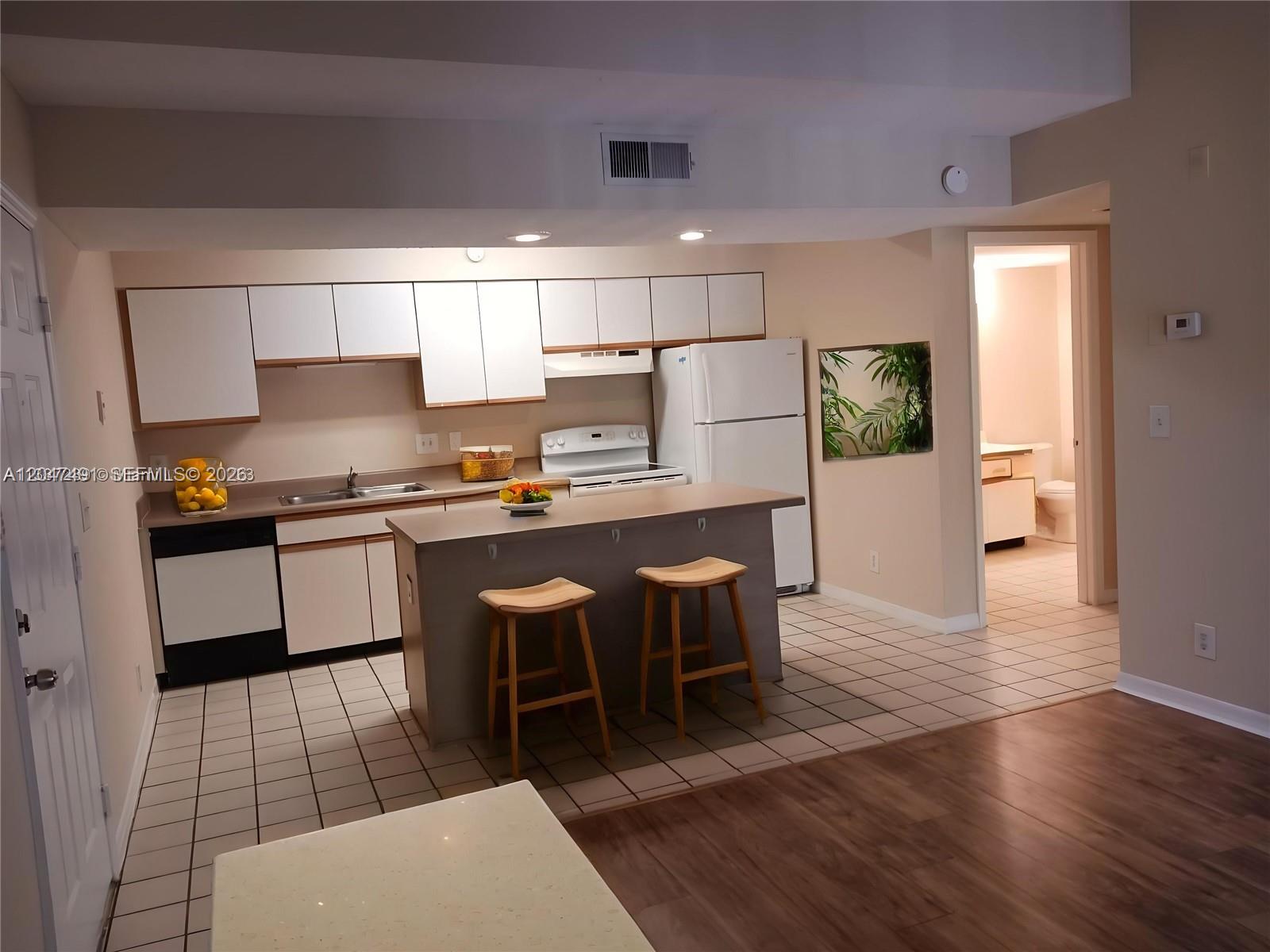 A kitchen features white cabinets, a central island with two stools, and tile flooring — POMPANO BEACH, FL