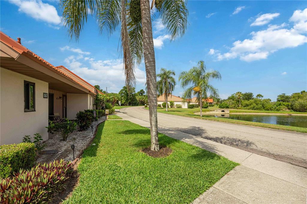 A single-story building with a red roof sits beside a grassy lawn and a pond — 6567 DRAW LANE UNIT 104, SARASOTA, FL