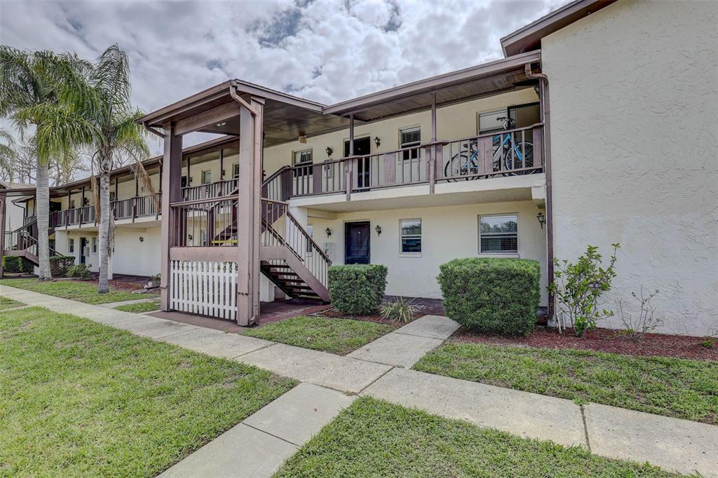 A two-story apartment building exterior features a wooden staircase, a balcony, and manicured landscaping — 8440 OLD POST ROAD UNIT 104, PORT RICHEY, FL