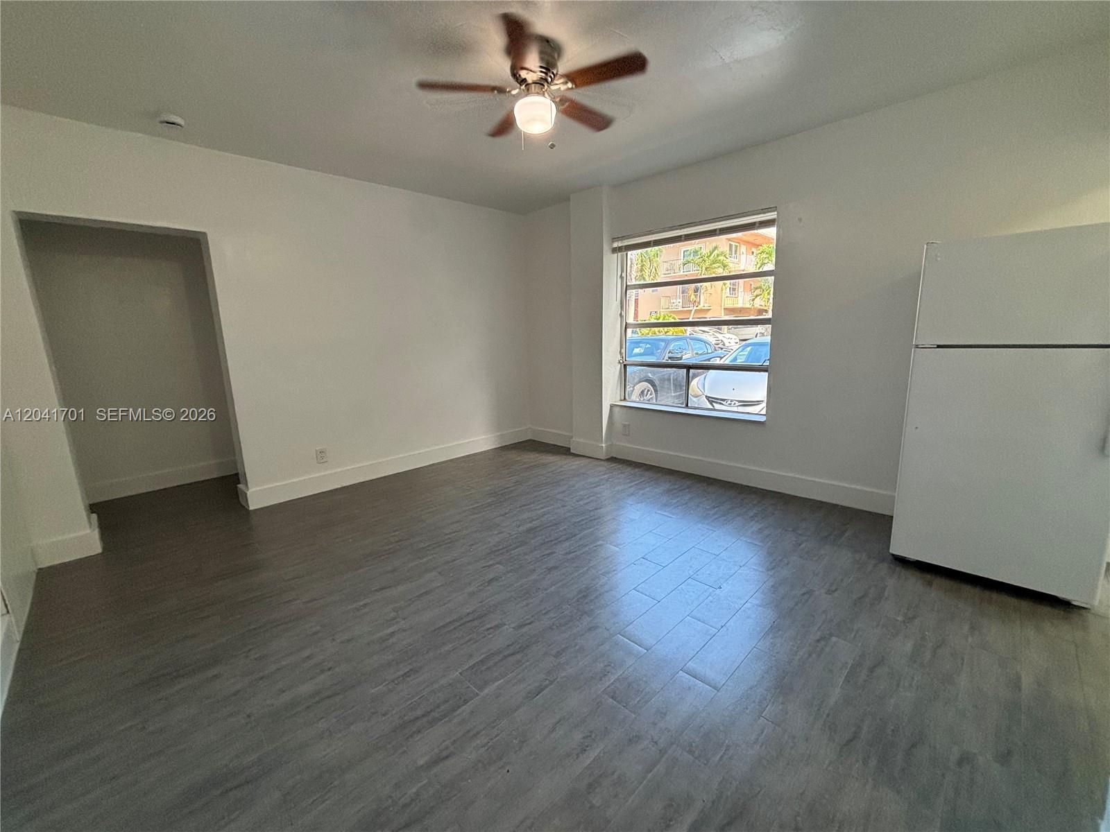 An empty room features gray wood-look flooring, white walls, a ceiling fan, and a refrigerator — 1843 Dewey St # 102, Hollywood FL 33020, HOLLYWOOD, FL