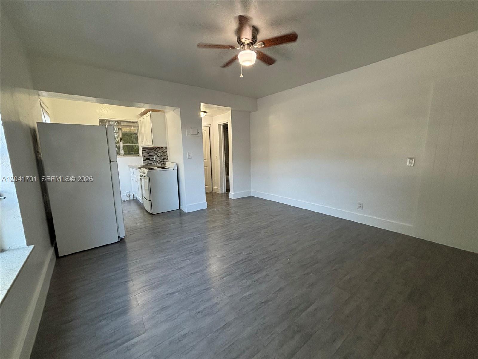 A living area with gray wood-look flooring leads into a small kitchen with white appliances — 1843 Dewey St # 102, Hollywood FL 33020, HOLLYWOOD, FL