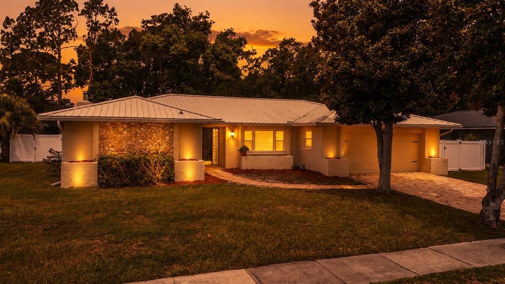A single-story house with a metal roof, stone facade, and illuminated landscaping at sunset — 8208 TANSY DRIVE, ORLANDO, FL