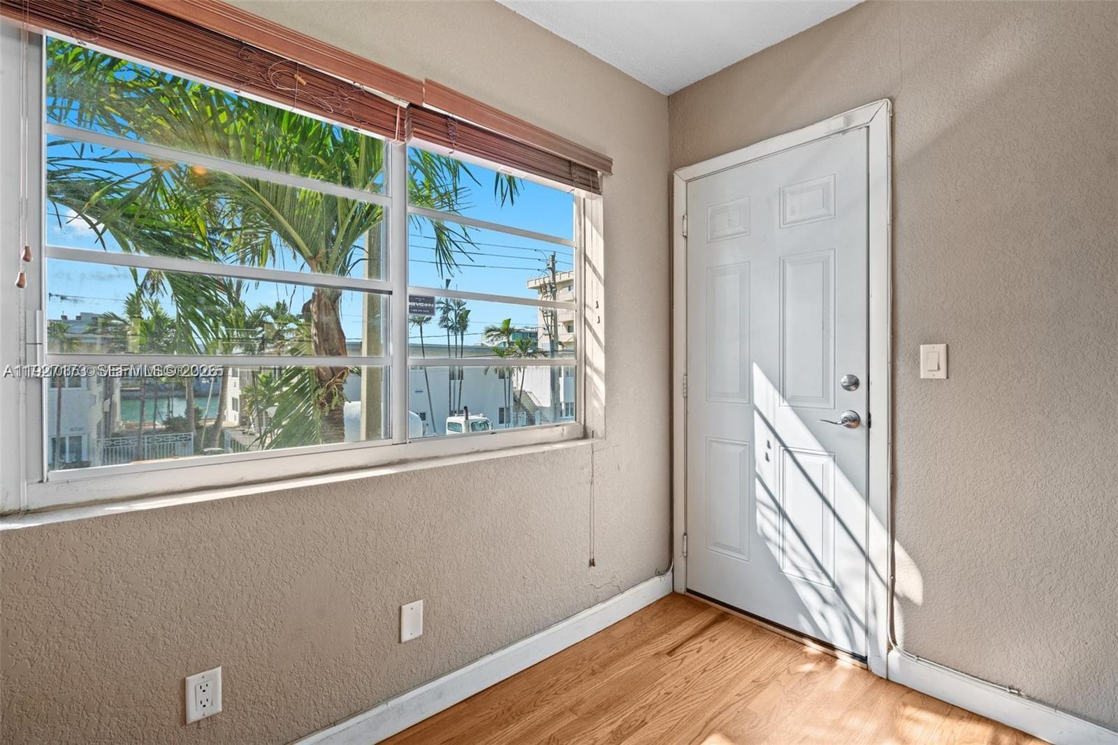 A room with hardwood floors, a white door, and a large window overlooking palm trees — 8345 Byron Ave # 6, Miami Beach FL 33141, MIAMI BEACH, FL