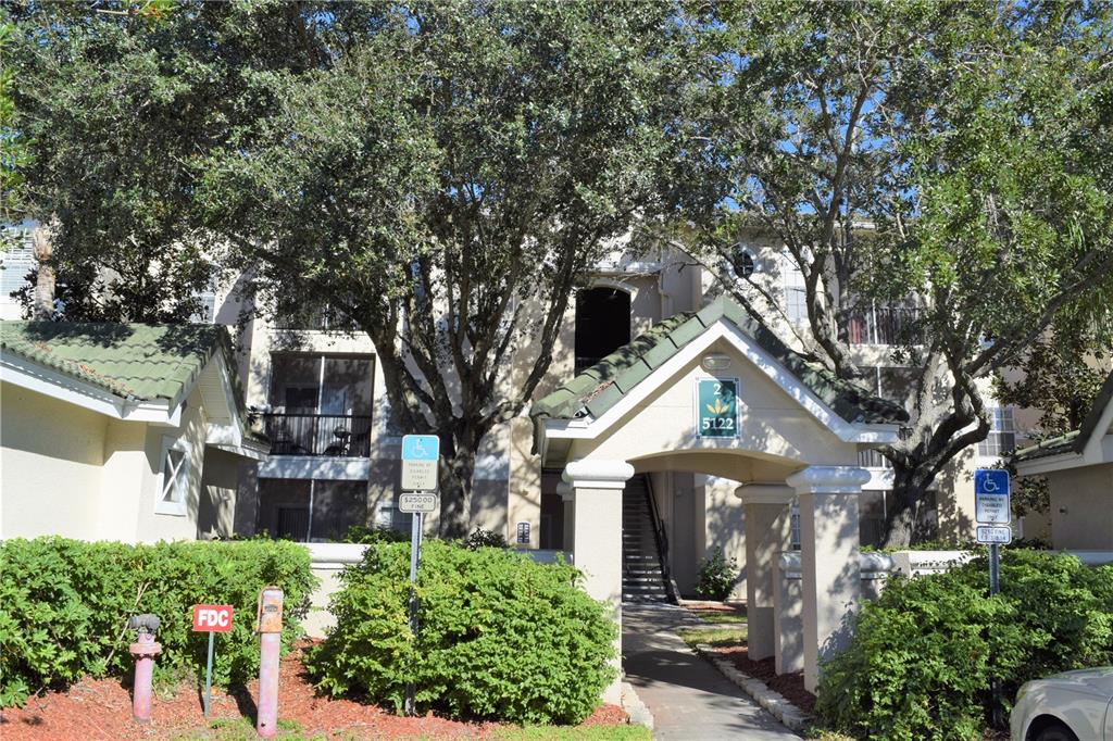 An exterior view of a multi-story apartment building entrance with a covered walkway and trees — 5122 NORTHRIDGE ROAD UNIT 202, SARASOTA, FL