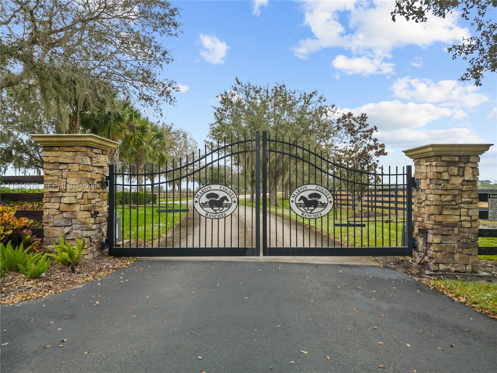 A black metal gate with circular emblems stands between two stone pillars at an entrance — 25750 SW Jockeys Run, Okeechobee FL 34974, OKEECHOBEE, FL