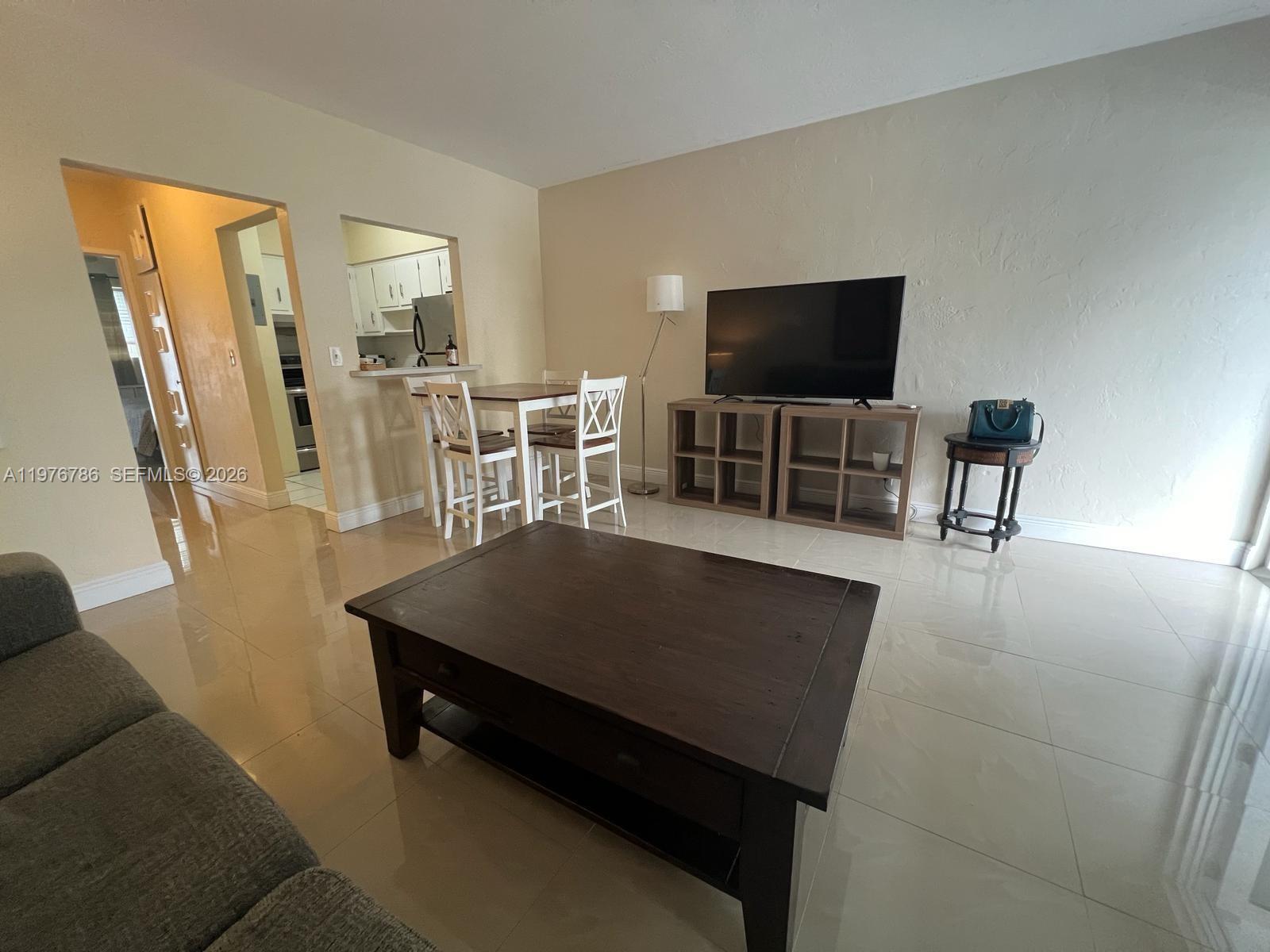 A living room features light-colored tile flooring, a dark coffee table, and a television stand — 600 Layne Blvd # 210, Hallandale Beach FL 33009, HALLANDALE BEACH, FL