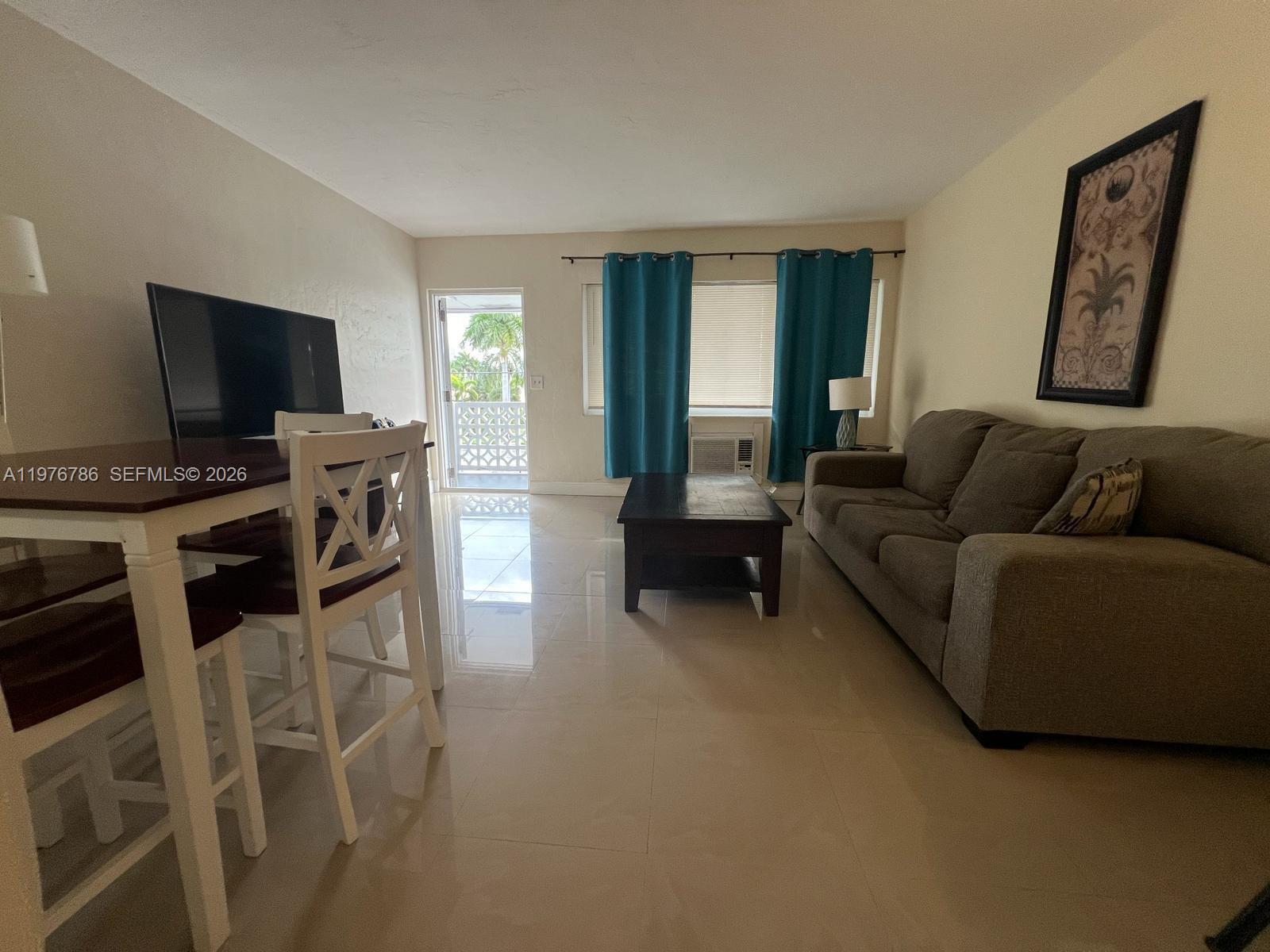 A living room features a sofa, coffee table, dining set, and a balcony door — 600 Layne Blvd # 210, Hallandale Beach FL 33009, HALLANDALE BEACH, FL