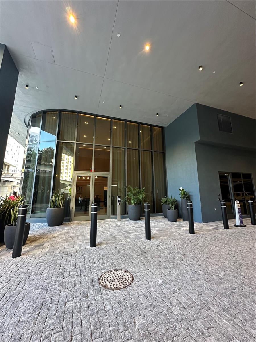 The exterior entrance of a modern building features glass doors, potted plants, and bollards — 121 NE 5 # 2712, Miami FL 33132, MIAMI, FL