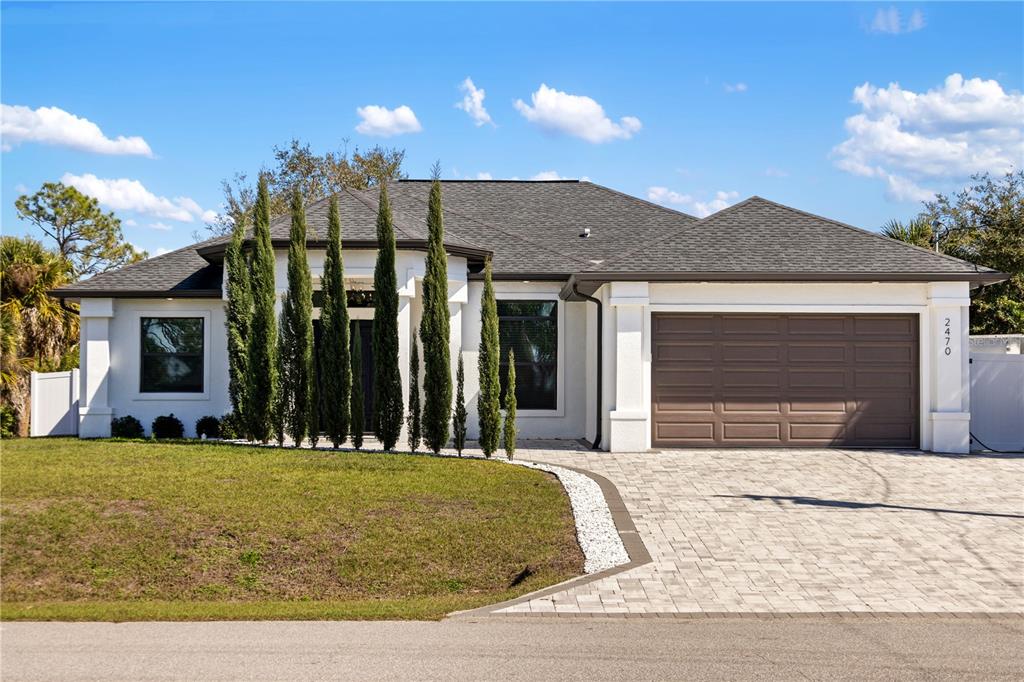 A single-story white house with a brown garage door and tall cypress trees in front — 2470 ABBOTSFORD STREET, NORTH PORT, FL
