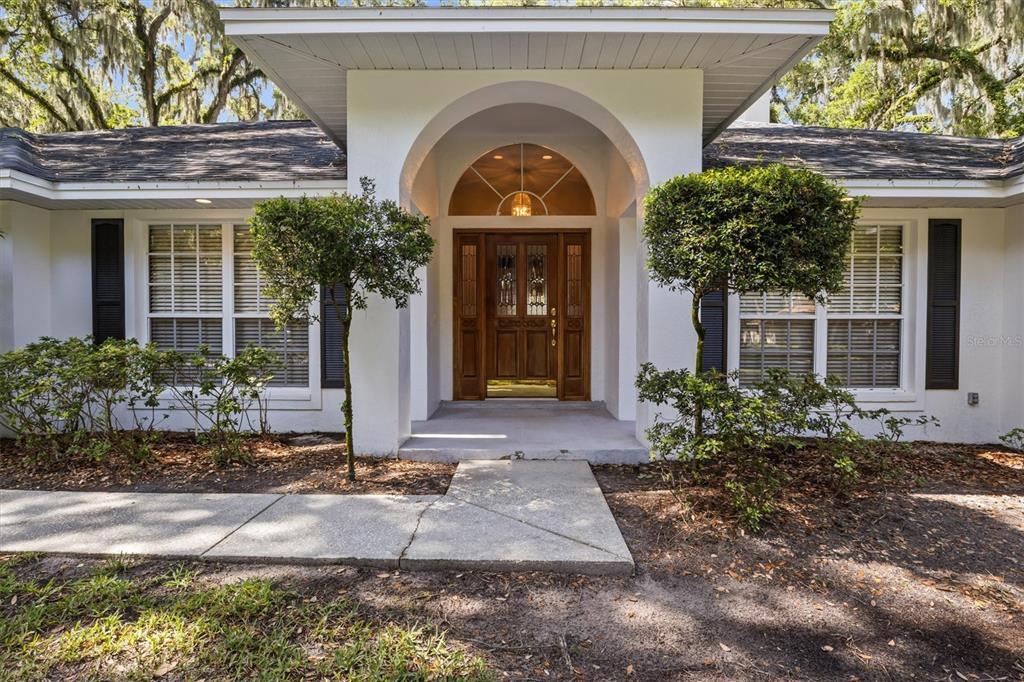 The front exterior of a white house features a prominent arched entryway and wooden doors — 1142 LONGWOOD OAKS BOULEVARD, LAKELAND, FL