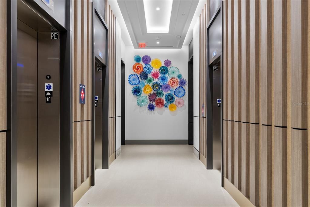 An elevator lobby features wood-paneled walls, tile flooring, and a colorful glass wall art installation — 275 1ST AVENUE S UNIT 1502, ST PETERSBURG, FL