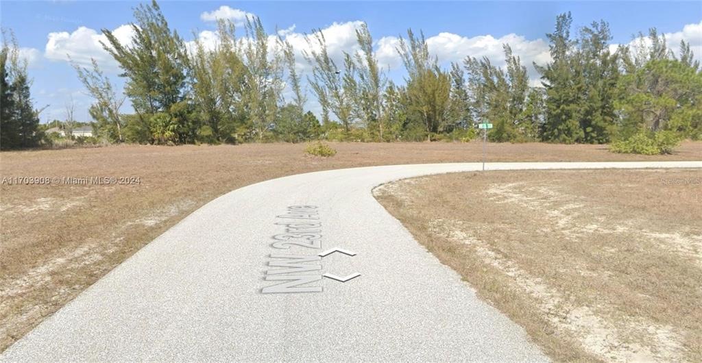 A paved road curves through a vacant, grassy lot with scattered trees under a blue sky — 1926 NW 23RD AVE AVENUE NW, CAPE CORAL, FL