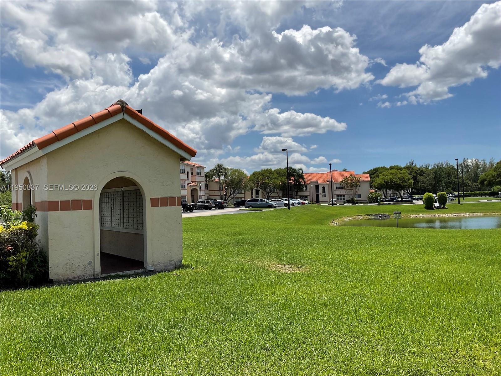 A small, tan mail kiosk with a red tile roof sits on a grassy lawn — 3320 N Pinewalk Dr N # 1715, Margate FL 33063, MARGATE, FL