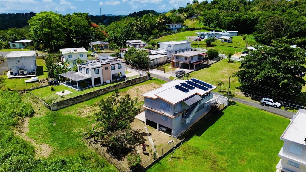 An aerial view shows several residential houses with solar panels surrounded by green, hilly terrain — Bo Ceiba Sur Sector Canal SOLAR #6, JUNCOS, PR