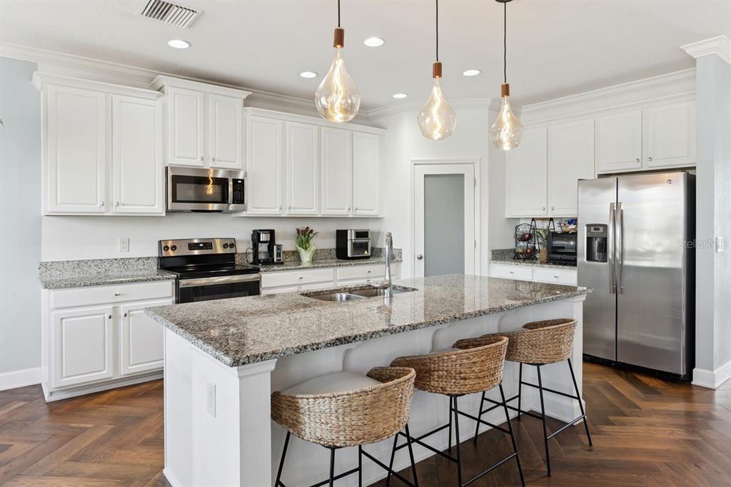 A kitchen features white cabinets, granite countertops, a central island with bar stools, and pendant lighting — 18799 HENEQUEN LANE SE, SPRING HILL, FL