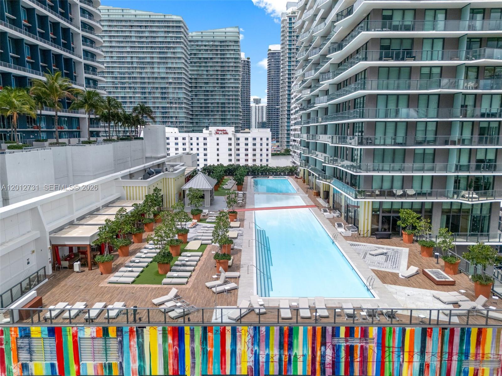 A large outdoor swimming pool is surrounded by lounge chairs, potted trees, and modern buildings — 1300 S Miami Ave, Miami FL 33130, MIAMI, FL
