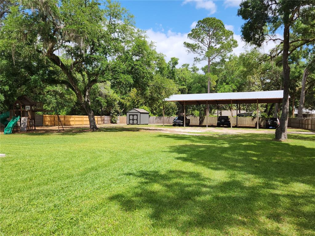 A large grassy backyard features a wooden playset, a storage shed, and a metal carport — 16189 SE 190TH AVENUE, WEIRSDALE, FL