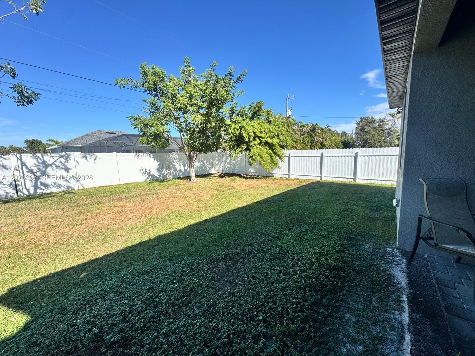 A grassy backyard features a white vinyl fence and a large tree under a blue sky — 209 SE 26th Ter, Cape Coral FL 33904, CAPE CORAL, FL
