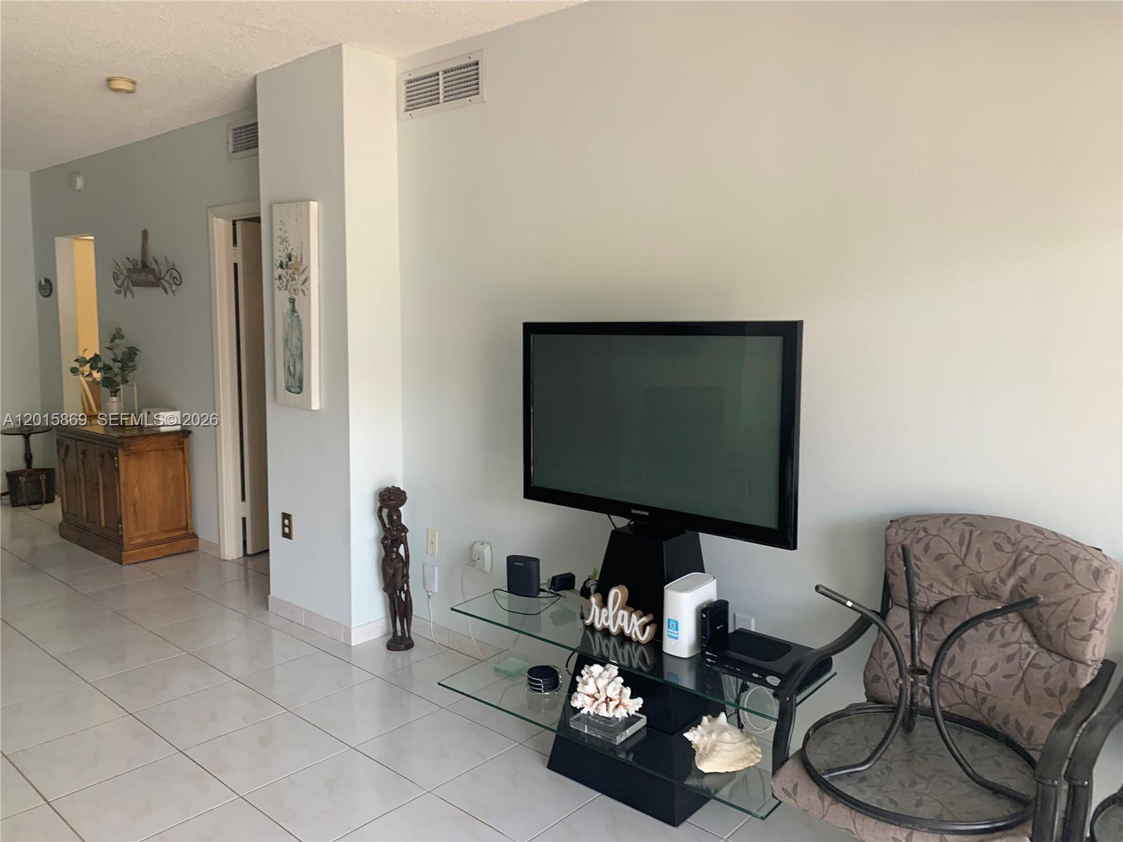 A living room features a television on a glass stand, tile flooring, and a wooden cabinet — 1985 S Ocean Dr, Hallandale Beach FL 33009, HALLANDALE BEACH, FL