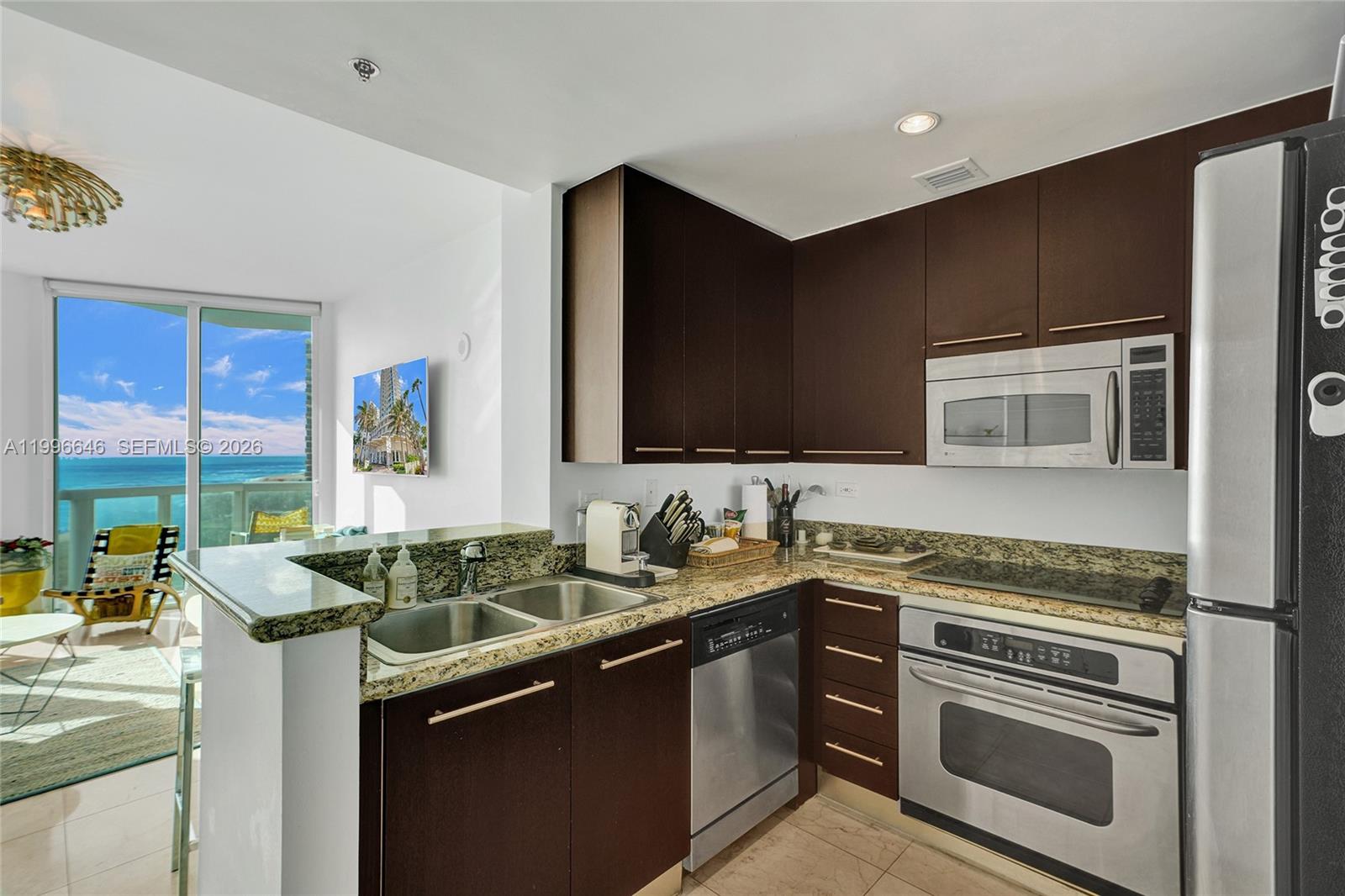 A kitchen with dark wood cabinets, granite countertops, and stainless steel appliances overlooks a balcony — 6515 Collins Ave # 804, Miami Beach FL 33141, MIAMI BEACH, FL