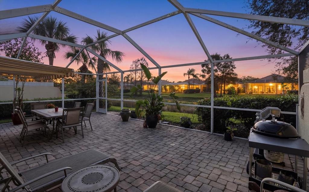 A screened-in patio features a paver floor, outdoor dining set, grill, and a sunset view — 5546 MODENA PLACE, SARASOTA, FL