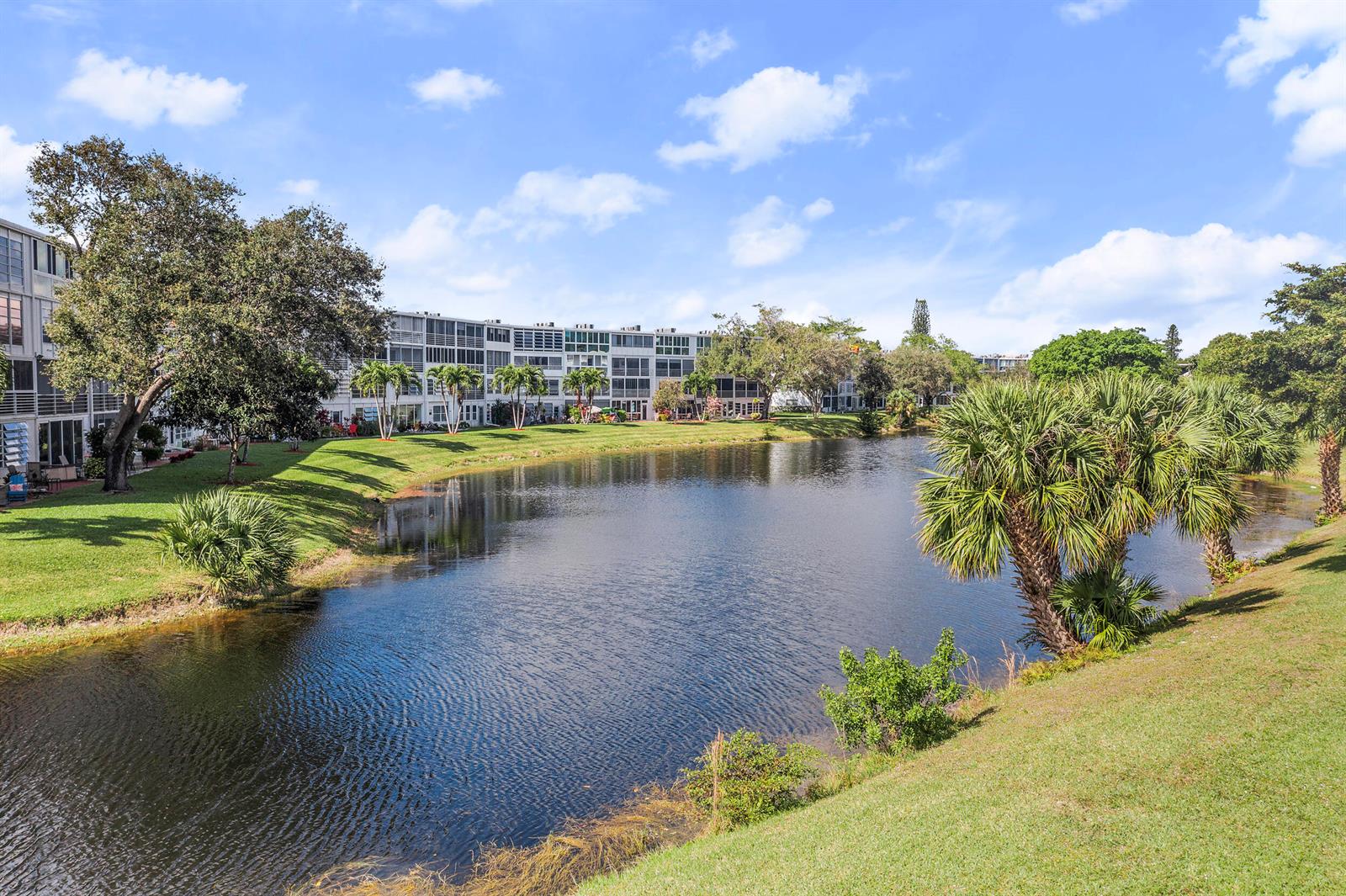 A multi-story apartment building sits behind a pond surrounded by green lawns and palm trees — 97 Oakridge H # 97, Deerfield Beach FL 33442, DEERFIELD BEACH, FL