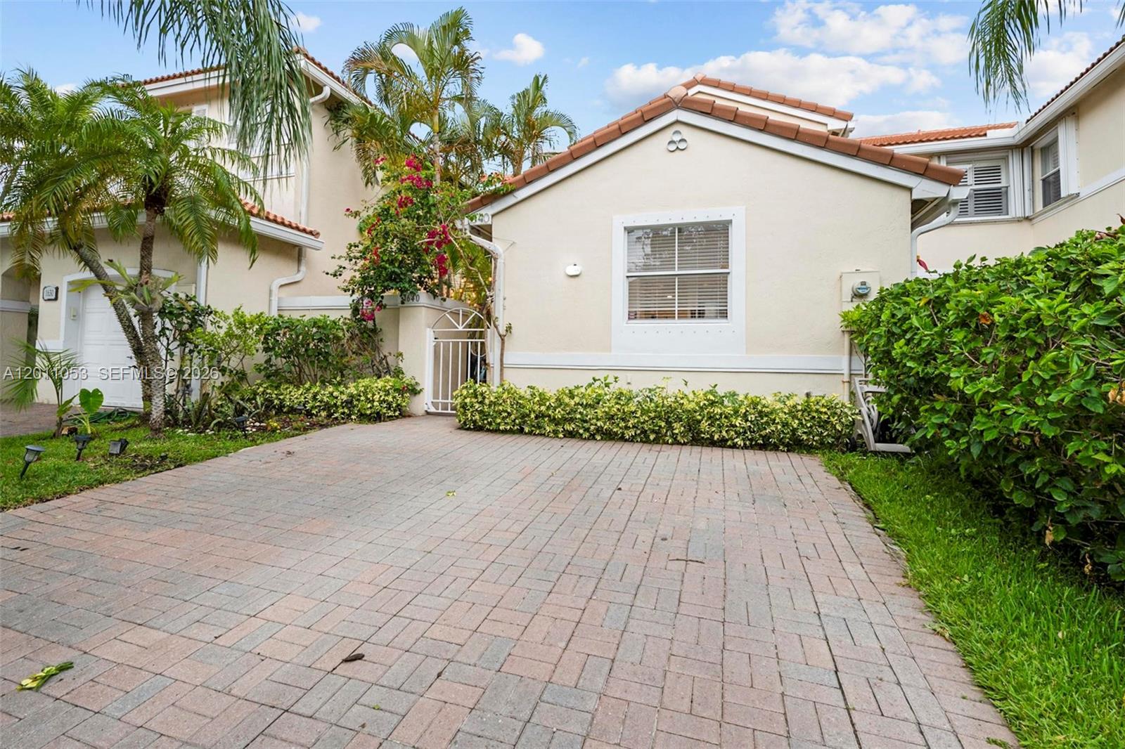 A paved driveway leads to a beige house with a white gate and landscaping — 1640 Weeping Willow Way # 1640, Hollywood FL 33019, HOLLYWOOD, FL