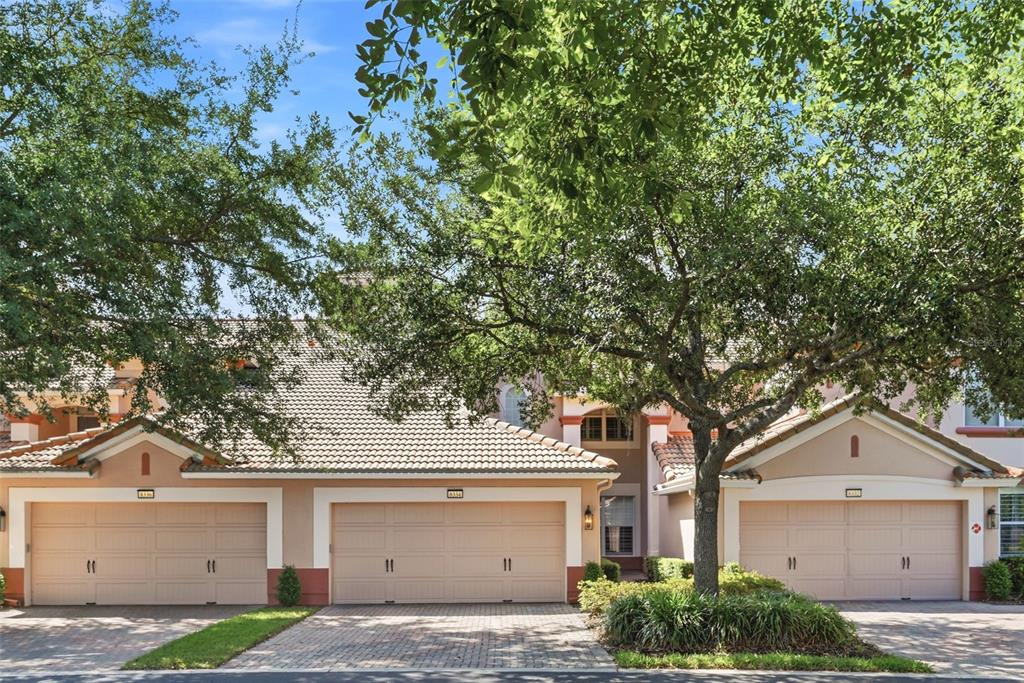 A three-car garage exterior of a tan stucco townhouse with a tiled roof and trees — 8334 RIVERDALE LANE UNIT 10320, DAVENPORT, FL