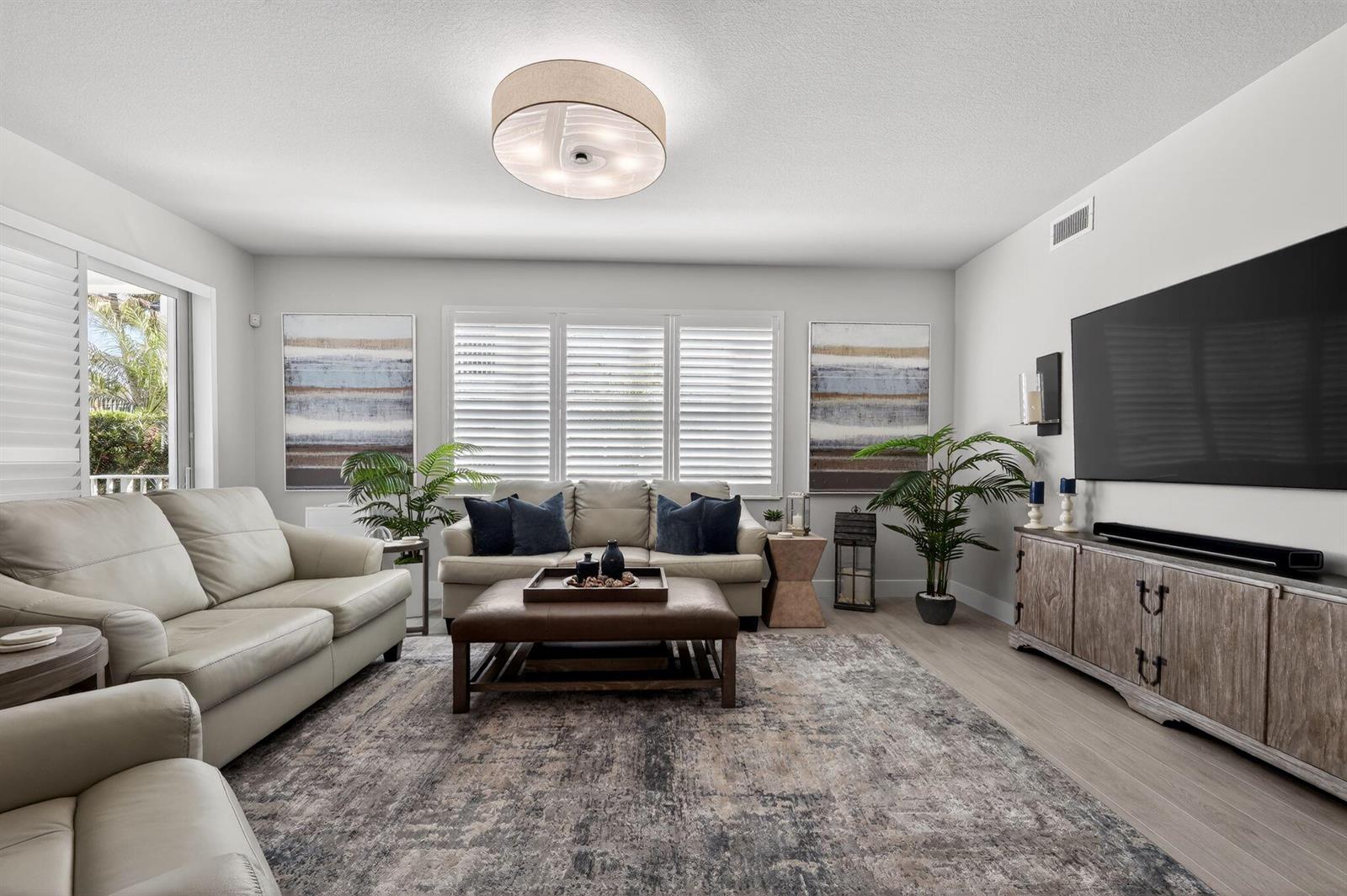 A living room features light-colored leather sofas, a central ottoman, and a large wall-mounted television — 915 Bay Colony Drive S, Juno Beach FL 33408, JUNO BEACH, FL