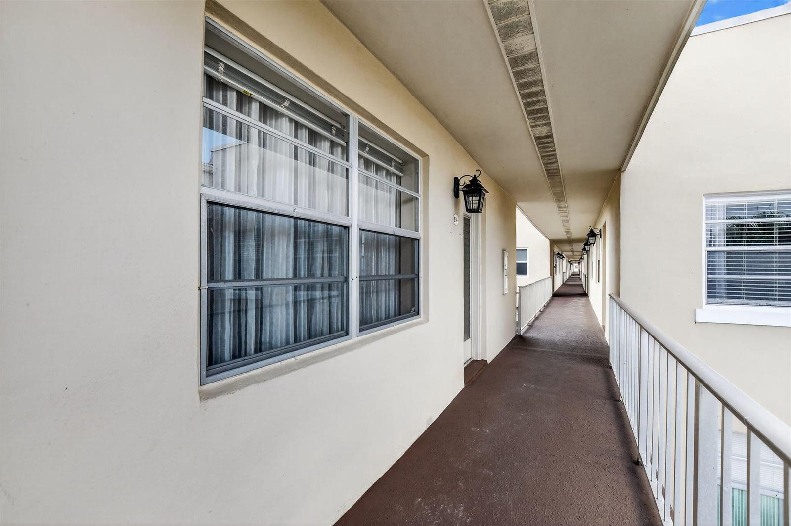 An exterior view of an apartment building walkway with a brown floor and white railings — 74 Burgundy B, Delray Beach FL 33484, DELRAY BEACH, FL