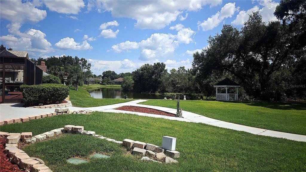 A grassy backyard features a stone-bordered garden, a concrete walkway, a gazebo, and a pond — 34826 CHELMSFORD LANE, ZEPHYRHILLS, FL