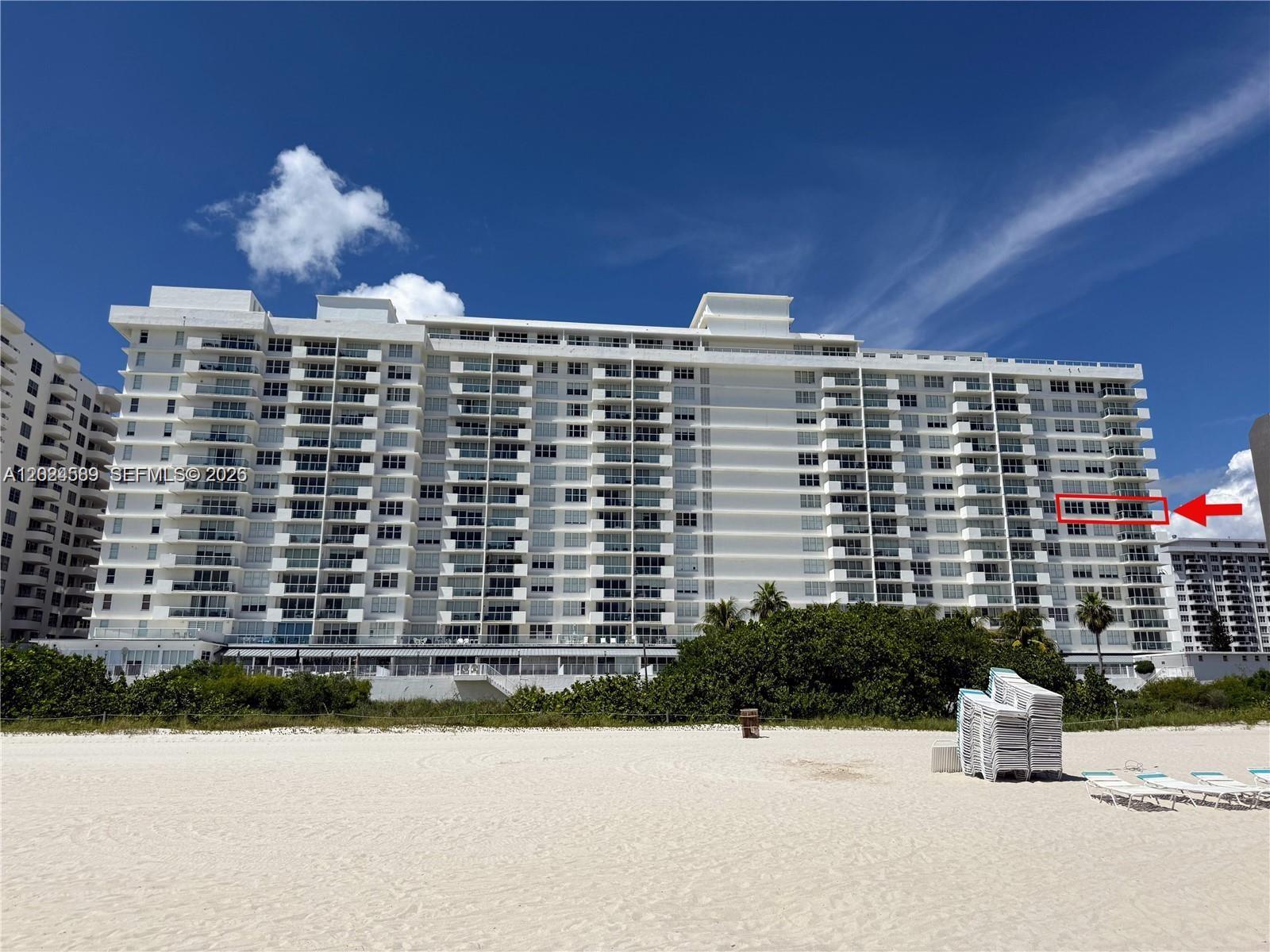 A multi-story white residential building stands behind a sandy beach under a blue sky — 5601 COLLINS AV # 607, Miami Beach FL 33140, MIAMI BEACH, FL