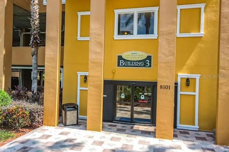 The exterior entrance of a yellow building features glass double doors and a trash receptacle — 8101 RESORT VILLAGE DRIVE UNIT 31501, ORLANDO, FL
