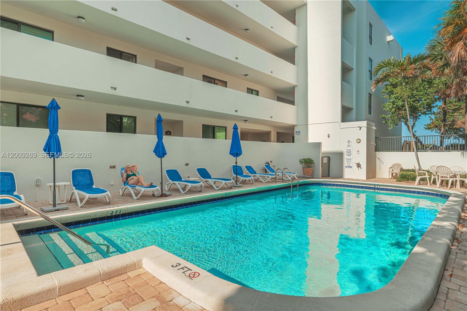 A swimming pool with lounge chairs and blue umbrellas sits beside a multi-story apartment building — 420 Celestial Way # 101, Juno Beach FL 33408, JUNO BEACH, FL