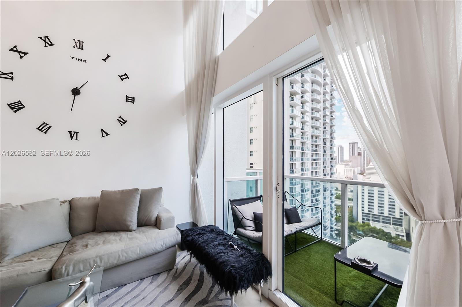 A living room features a large wall clock, a grey sofa, and a balcony view — 41 SE 5th St # 1303, Miami FL 33131, MIAMI, FL
