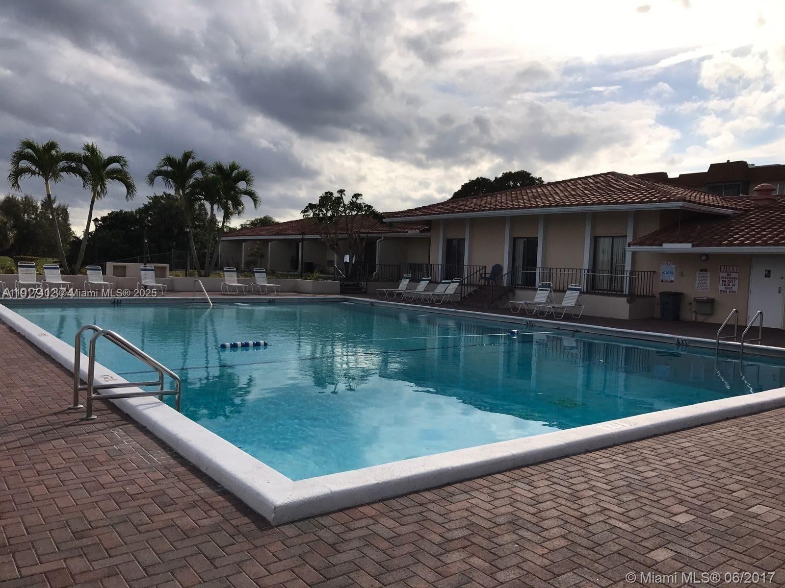 A rectangular swimming pool with a brick deck and lounge chairs sits before a building — 4174 Inverrary Dr # 1004, Lauderhill FL 33319, LAUDERHILL, FL