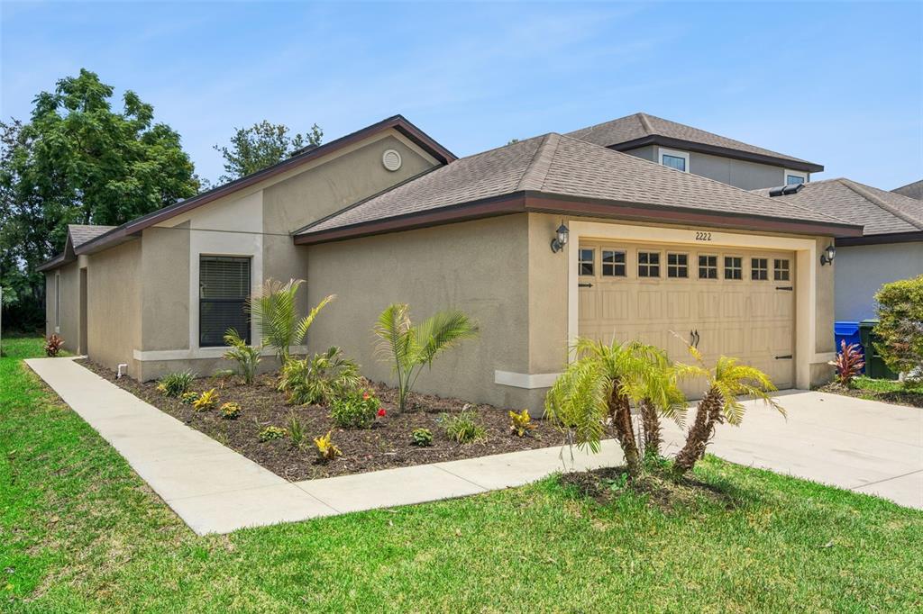 A tan single-story house features a two-car garage, a concrete walkway, and landscaped front yard — 2222 CASPIAN DRIVE, LAKELAND, FL