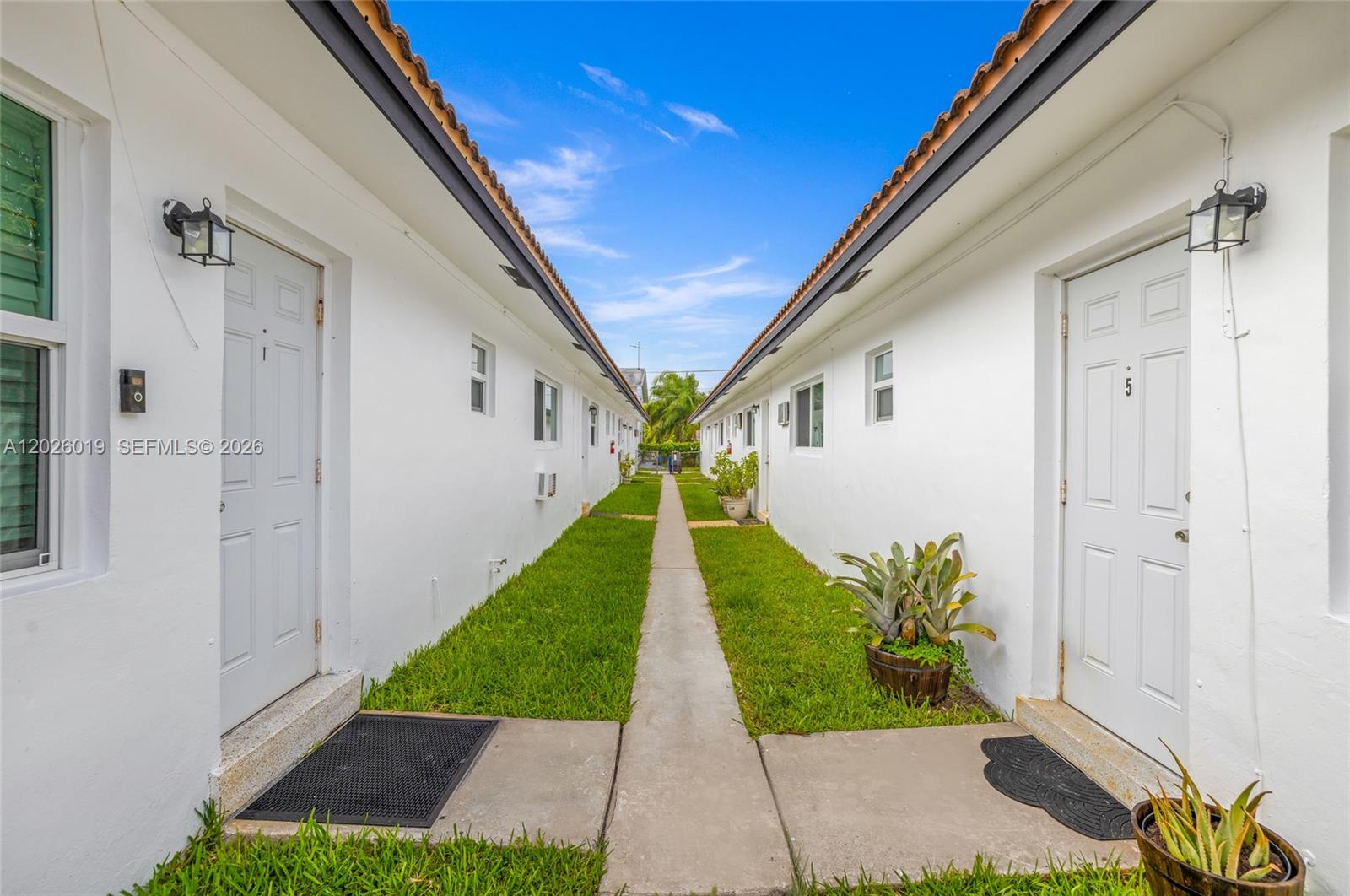 A narrow concrete walkway separates two rows of white apartment units with green grass lawns — 1927/1929 Roosevelt St, Hollywood FL 33020, HOLLYWOOD, FL