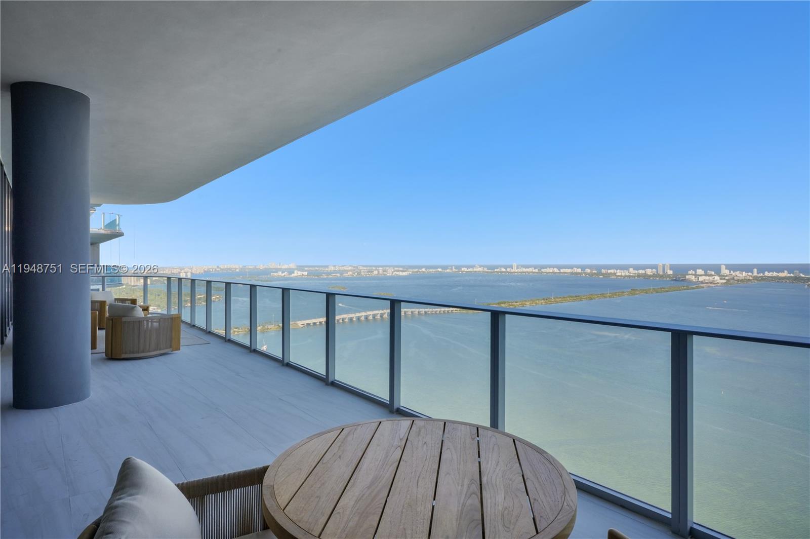 A high-rise balcony features a glass railing, wooden furniture, and a panoramic bay view — 700 NE 24, Miami FL 33137, MIAMI, FL