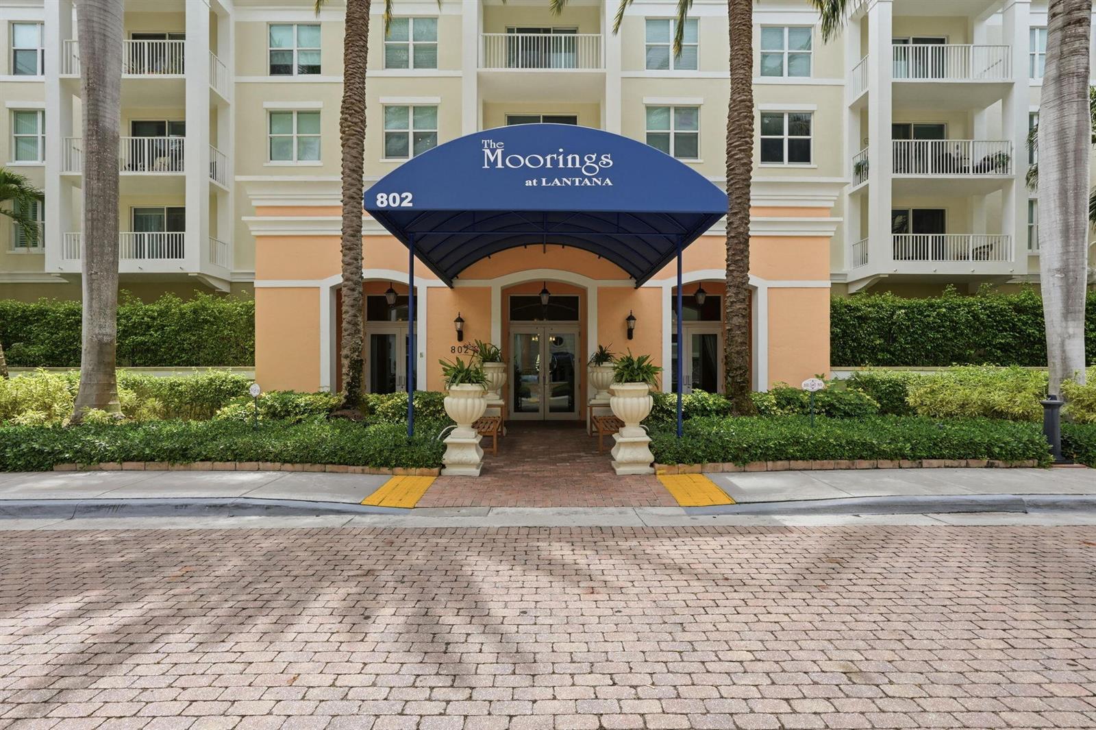The exterior entrance of a multi-story apartment building features a blue awning and palm trees — 802 W Windward Way # 304, Lantana FL 33462, LANTANA, FL