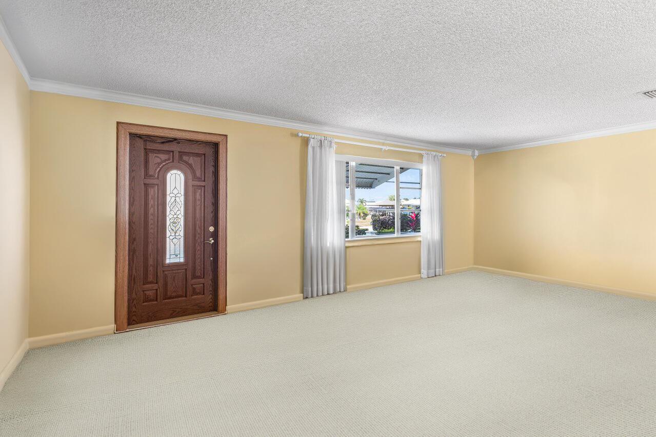 An empty room with beige walls, light carpet, a wooden front door, and a window — 1802 SW 13th Avenue, Boynton Beach FL 33426, BOYNTON BEACH, FL