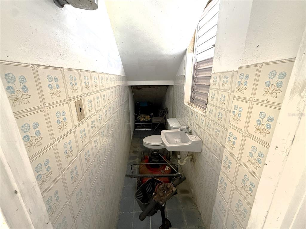 A narrow, tiled bathroom contains a toilet, sink, and miscellaneous items under a sloped ceiling — 12 HOSTOS CABRERA ST., SAN SEBASTIAN, PR