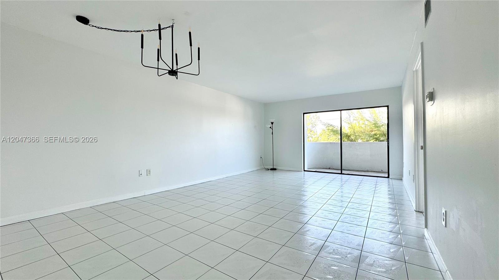 An empty room features white walls, light-colored tile flooring, a modern chandelier, and a sliding glass door — 130 S Shore Dr # 4B, Miami Beach FL 33141, MIAMI BEACH, FL
