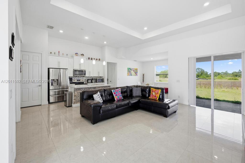 A living room with a black sectional sofa opens to a kitchen and backyard view — 1211 Eclat St E, Lehigh Acres FL 33974, LEHIGH ACRES, FL